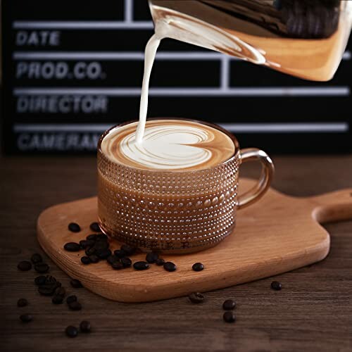 Latte art being poured into a textured glass mug on a wooden board with coffee beans.