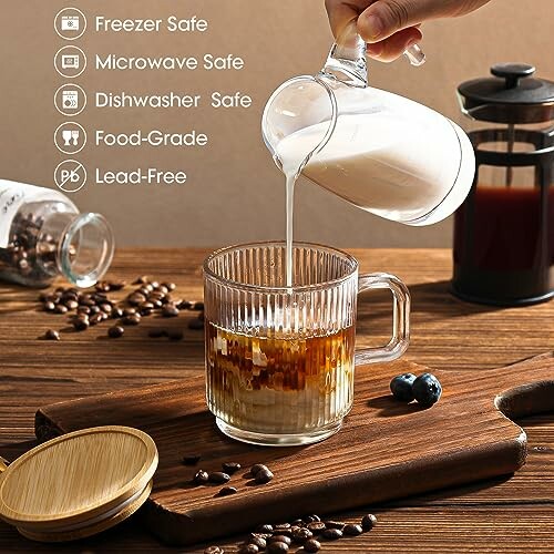 Glass mug of coffee with milk being poured, surrounded by coffee beans and French press.